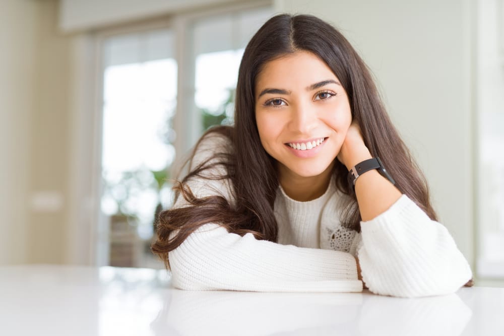 Smiling young woman leaning on table in bright home - Dentist Nocatee Young woman with long dark hair wearing a white sweater, smiling warmly while leaning on a table in a bright, modern room - Dentist Nocatee