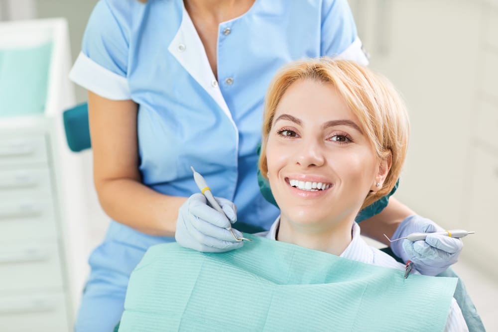 Blonde woman smiling while sitting in a dental chair with a bib on, as a dental professional in scrubs prepares tools for treatment - Zoom Teeth Whitening