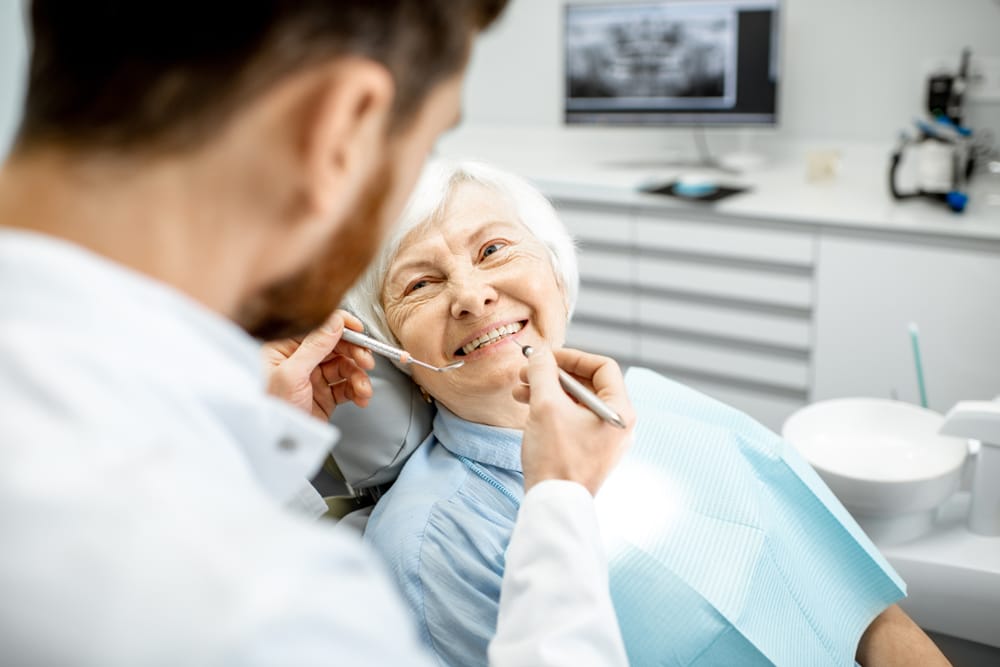 Smiling Senior Patient During Jaw Exam – TMJ Treatment Older female patient smiling as a dentist examines her jaw and bite alignment in a modern dental office – TMJ Treatment