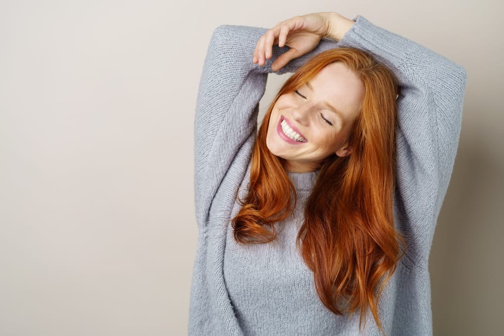 Joyful red-haired woman smiling with eyes closed - Teeth Whitening St Augustine Cheerful woman with long red hair wearing a cozy gray sweater, smiling brightly with her eyes closed and arms raised - Teeth Whitening St Augustine