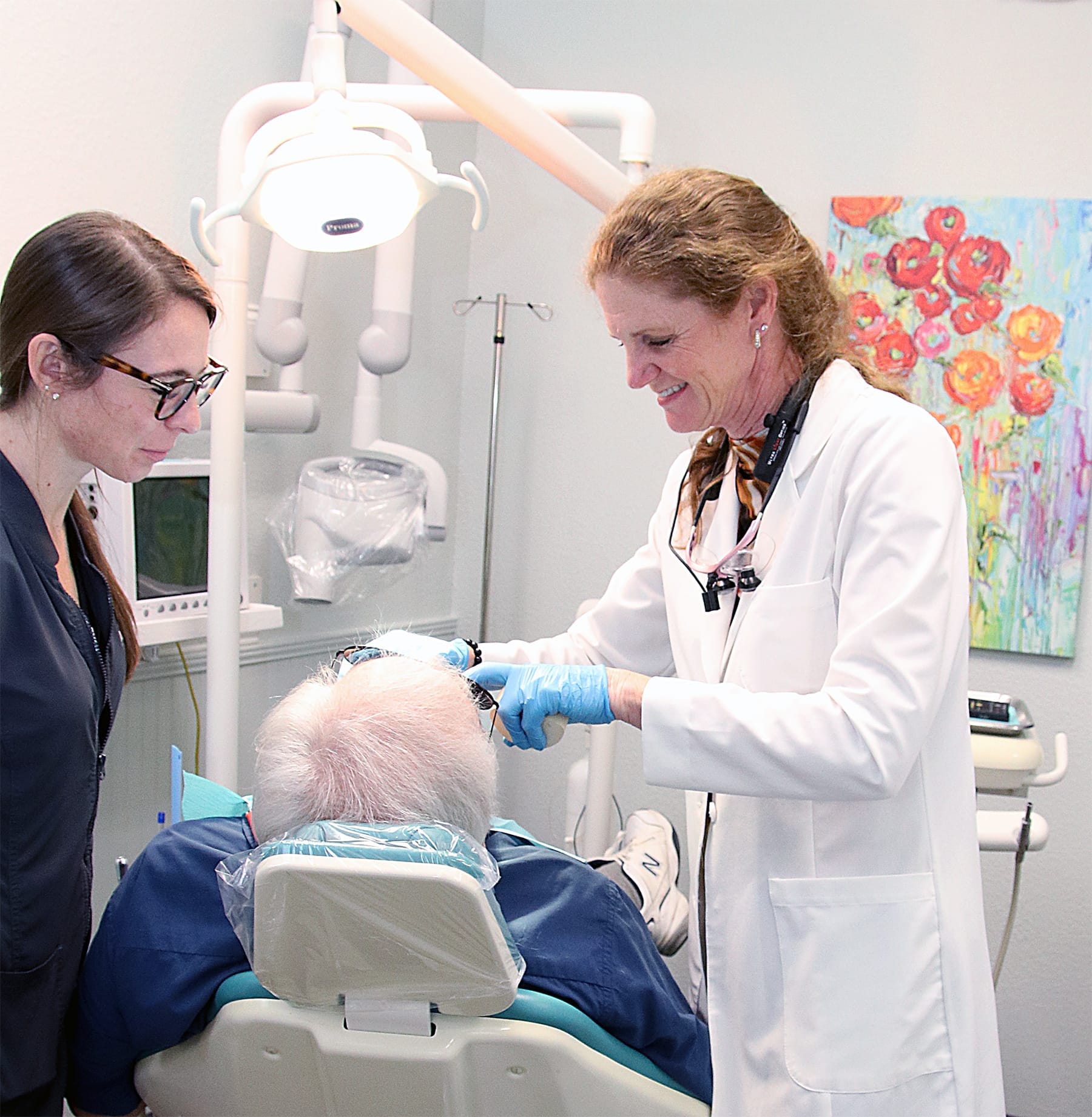 Professional Dental Consultation with Patient – Cosmetic Dentistry  A smiling female dentist in a white coat and her assistant consulting with an elderly male patient in a dental chair, surrounded by modern equipment and a colorful flower painting – Cosmetic Dentistry