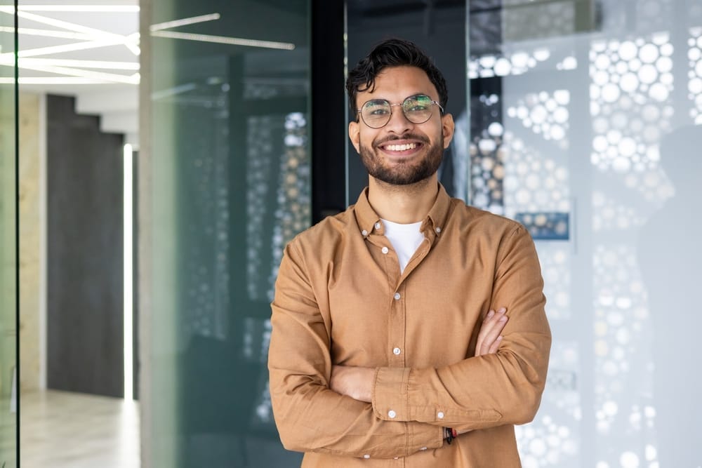 Confident Man Smiling with Arms Crossed – Cosmetic Dentistry St Augustine A young man wearing glasses and a brown shirt smiles confidently with his arms crossed, standing in a modern office environment – Cosmetic Dentistry St Augustine