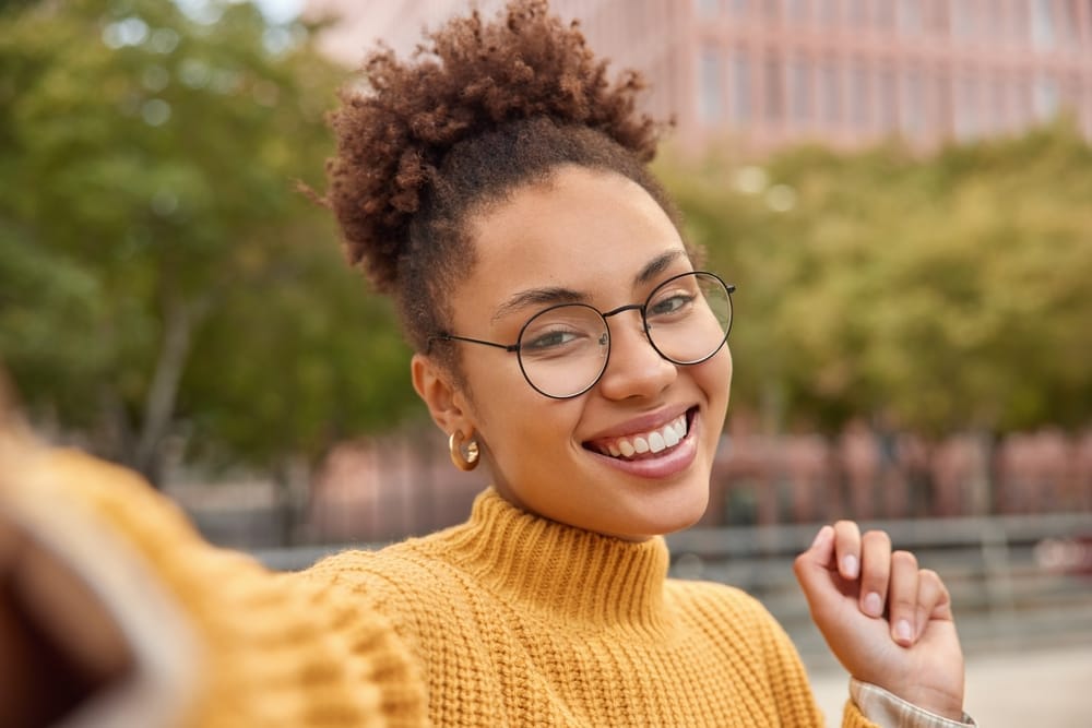 Happy young woman taking a selfie outdoors in a yellow sweater - Dental Bridge St Augustine Smiling young woman with glasses and curly hair wearing a yellow turtleneck sweater, taking a selfie outside in an urban park - Dental Bridge St Augustine