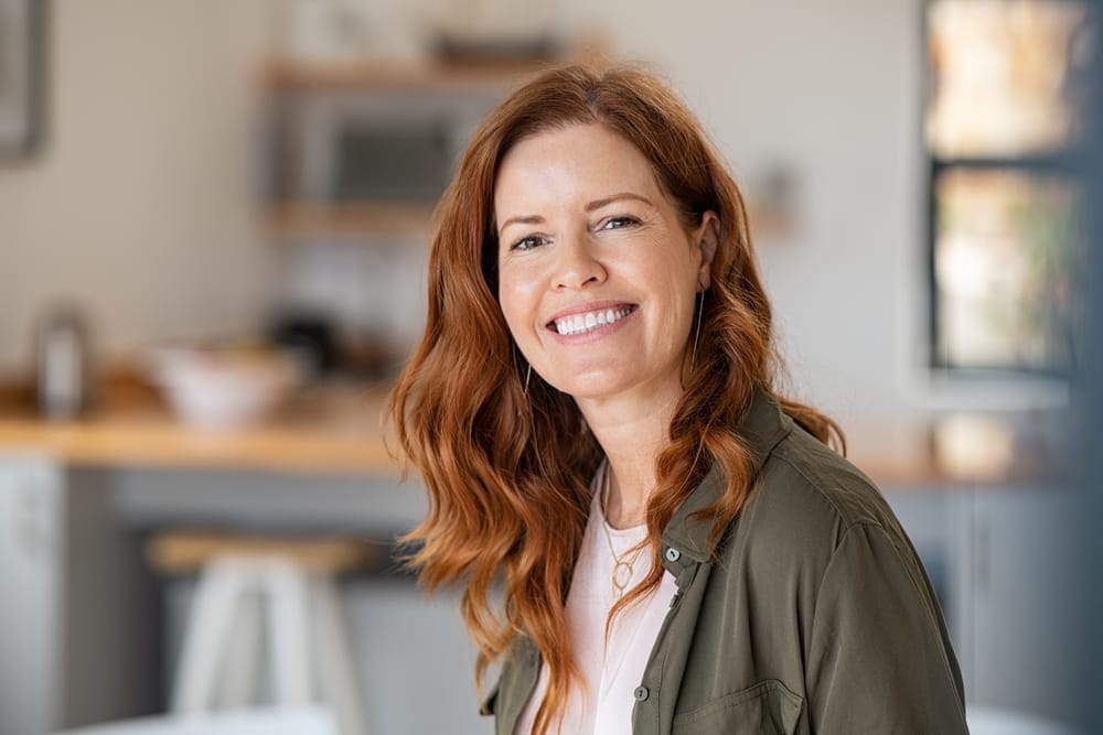 Mature woman with red hair smiling at home - Dental Implants St Augustine Portrait of a smiling woman with long red hair wearing a casual green jacket, sitting in a bright and modern home environment - Dental Implants St Augustine