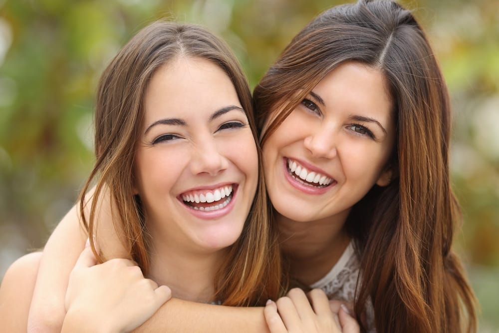 Close-up of two young women with long hair hugging and laughing together outside, showing off their bright smiles - Veneers St Augustine