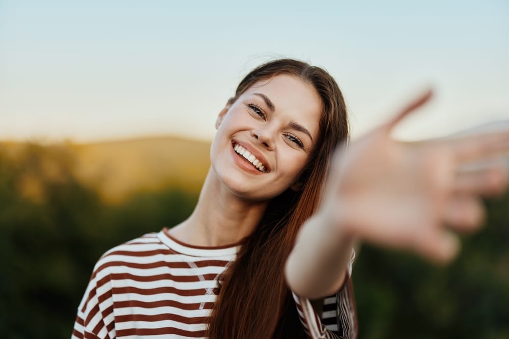 Cheerful young woman with long brown hair wearing a striped shirt, smiling widely and extending her arm toward the camera in a natural outdoor setting - Dentures St Augustine