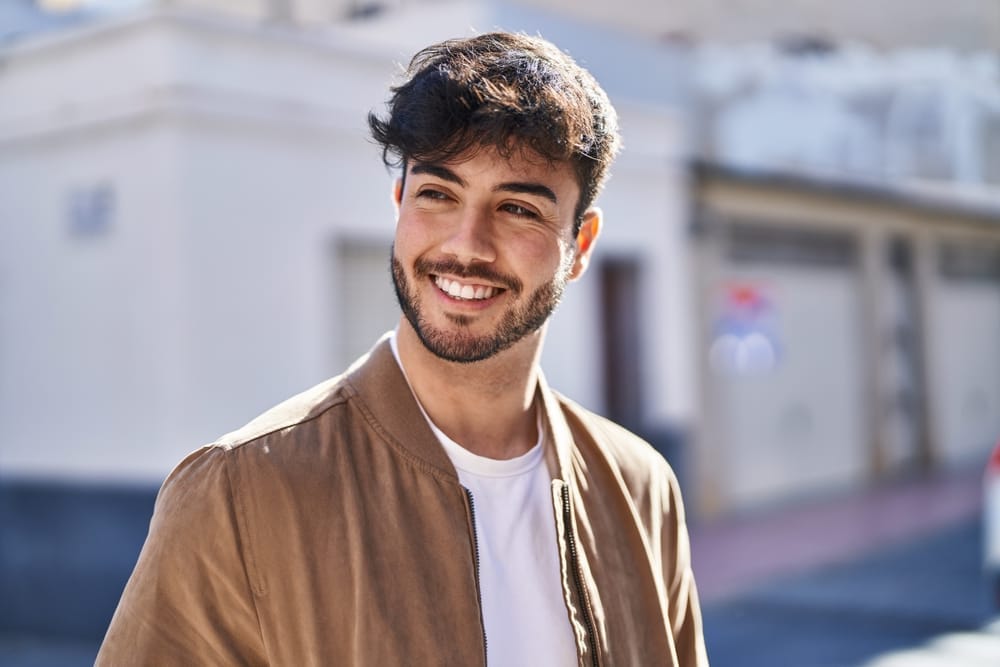 Young man smiling confidently outdoors in casual jacket - Dental Crowns St Augustine Portrait of a smiling young man with dark hair and a beard, wearing a tan jacket and standing outside in a sunny urban setting - Dental Crowns St Augustine
