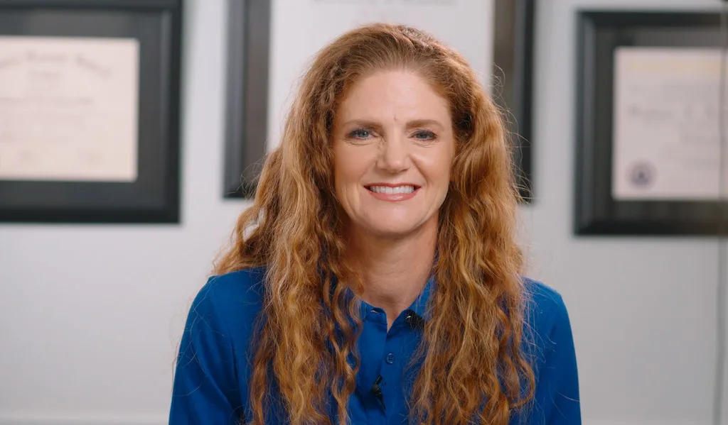 Dr. Stephanie Kinsey Smiling Indoors – Palencia Dental Dr. Stephanie Kinsey, wearing a blue blouse, smiles warmly while seated indoors with framed certificates in the background – Palencia Dental