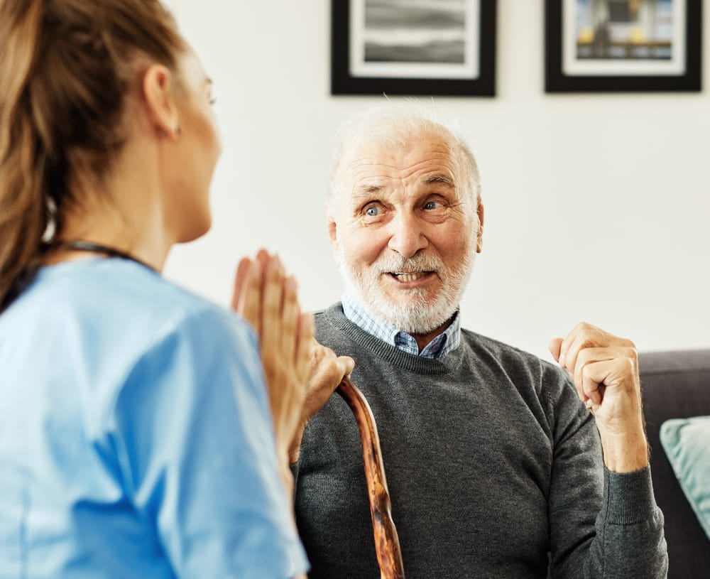 Elderly Man Smiling During Care Consultation – Orthodontics Happy senior man with a cane interacting with a healthcare professional in scrubs, showing improved confidence and alignment – Orthodontics
