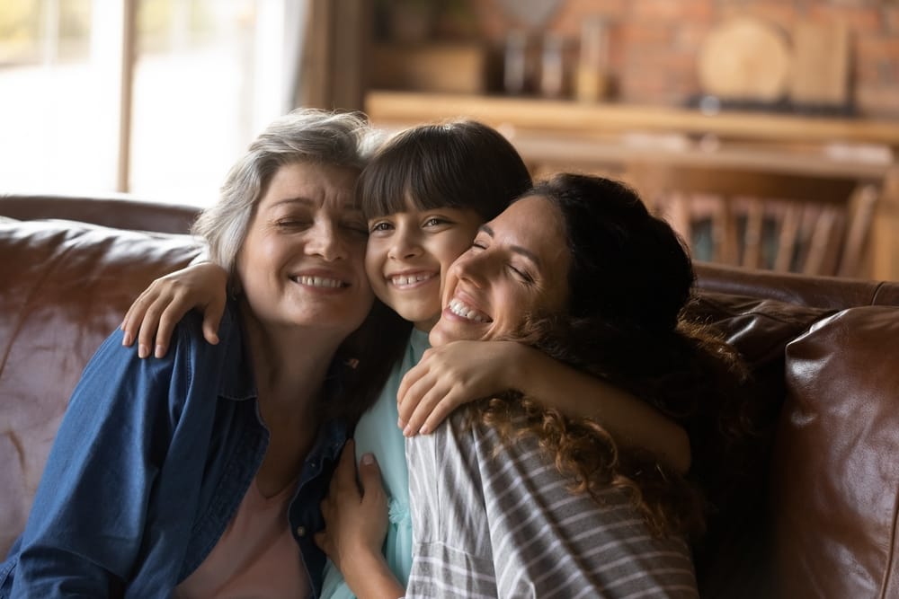 Happy Family Hugging on Couch – Orthodontics Three generations of a smiling family, including a young girl, her mother, and grandmother, share a warm hug while sitting on a couch at home – Orthodontics