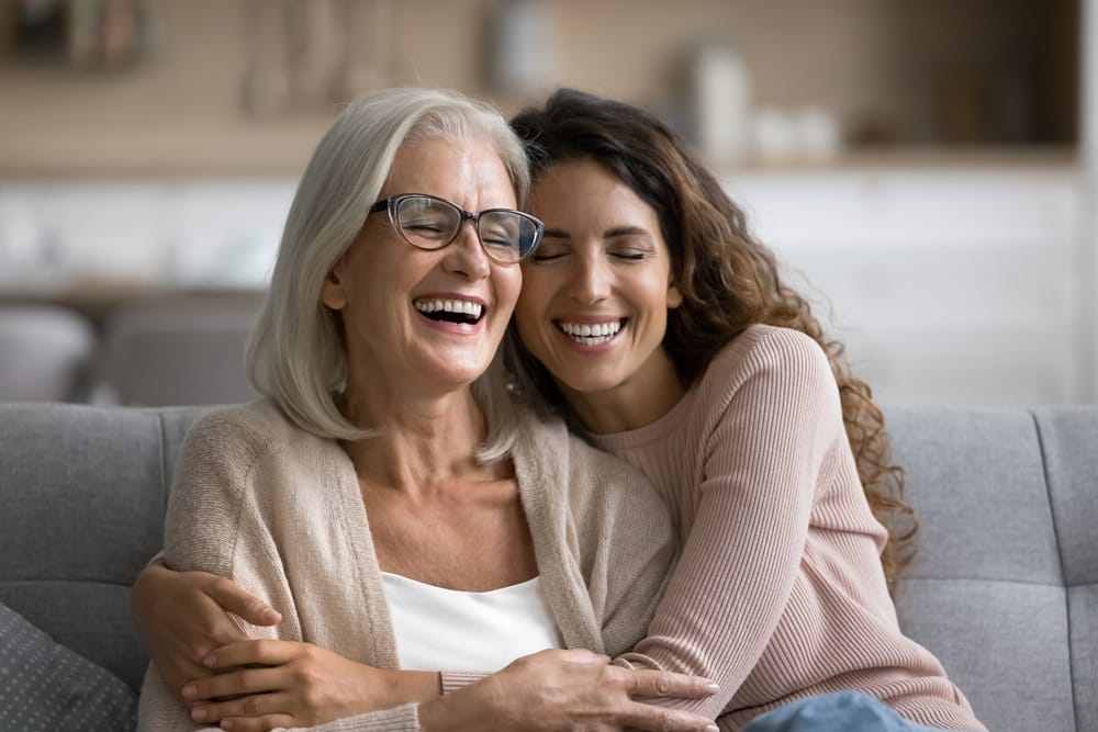 Happy Mother and Daughter Hugging on Couch – Dentist St Augustine FL An older woman with gray hair and glasses hugs and laughs with her adult daughter, both smiling warmly while sitting on a couch – Dentist St Augustine FL
