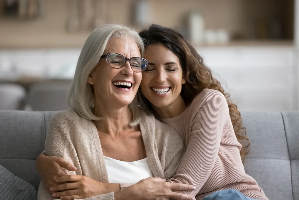 An older woman with gray hair and glasses hugs and laughs with her adult daughter, both smiling warmly while sitting on a couch – Dentist St Augustine FL