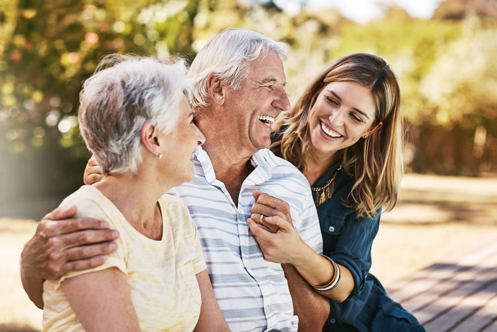 Happy older couple sitting on a bench with their adult daughter, all smiling and laughing together in a sunny outdoor park setting - TMJ Treatment