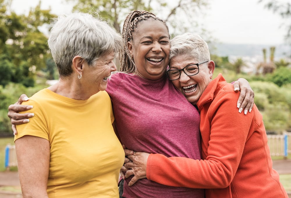 Three older women smiling and hugging while laughing outdoors in a park setting, enjoying a pain-free moment – Headaches 