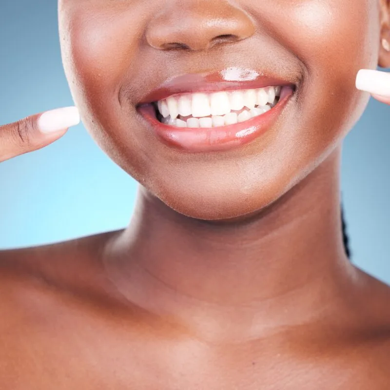 Close-up of a woman pointing to her radiant, healthy smile with white teeth against a blue background, representing confidence, beauty, and the power of your smile
