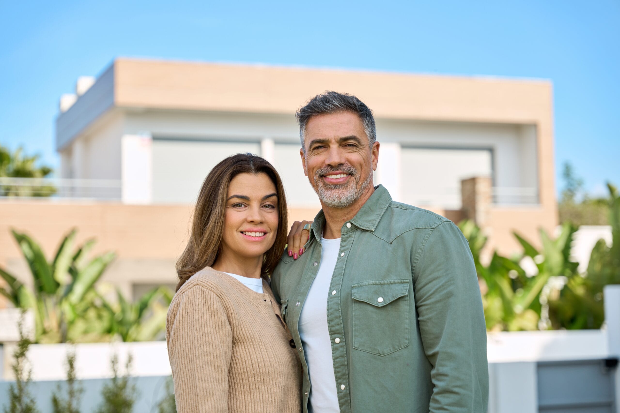 Smiling middle-aged couple standing outside modern home - Cosmetic Dentistry Happy middle-aged couple posing outdoors in front of a contemporary house, both smiling and dressed casually with tropical greenery in the background - Cosmetic Dentistry